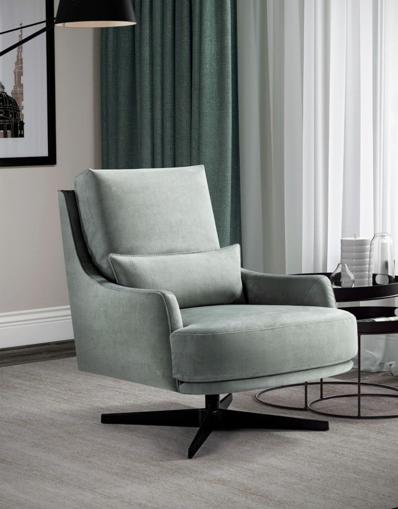 Filicudi armchair in pale sage fabric on a black swivel base, three-quarter view in a living room with teal curtains
