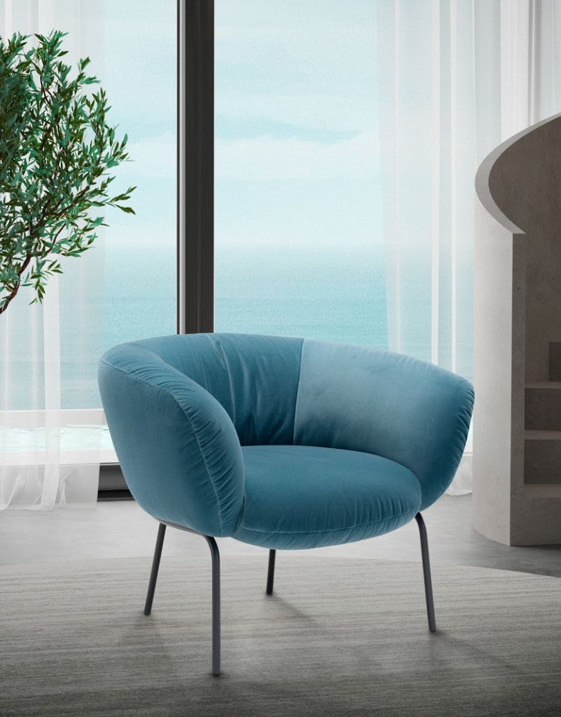 Jill armchair in blue velvet with slim dark metal legs in a bright living room overlooking the sea